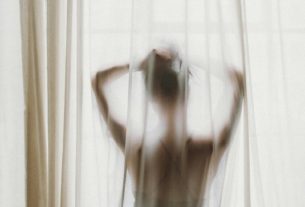 A person in silhouette behind a sheer white curtain, hands raised to their hair