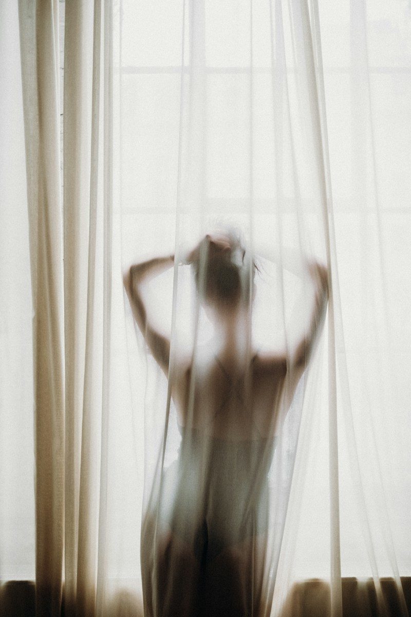 A person in silhouette behind a sheer white curtain, hands raised to their hair